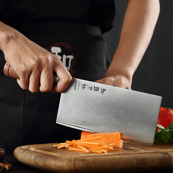 Person using a large knife to slice carrots on a wooden cutting board with a dark background